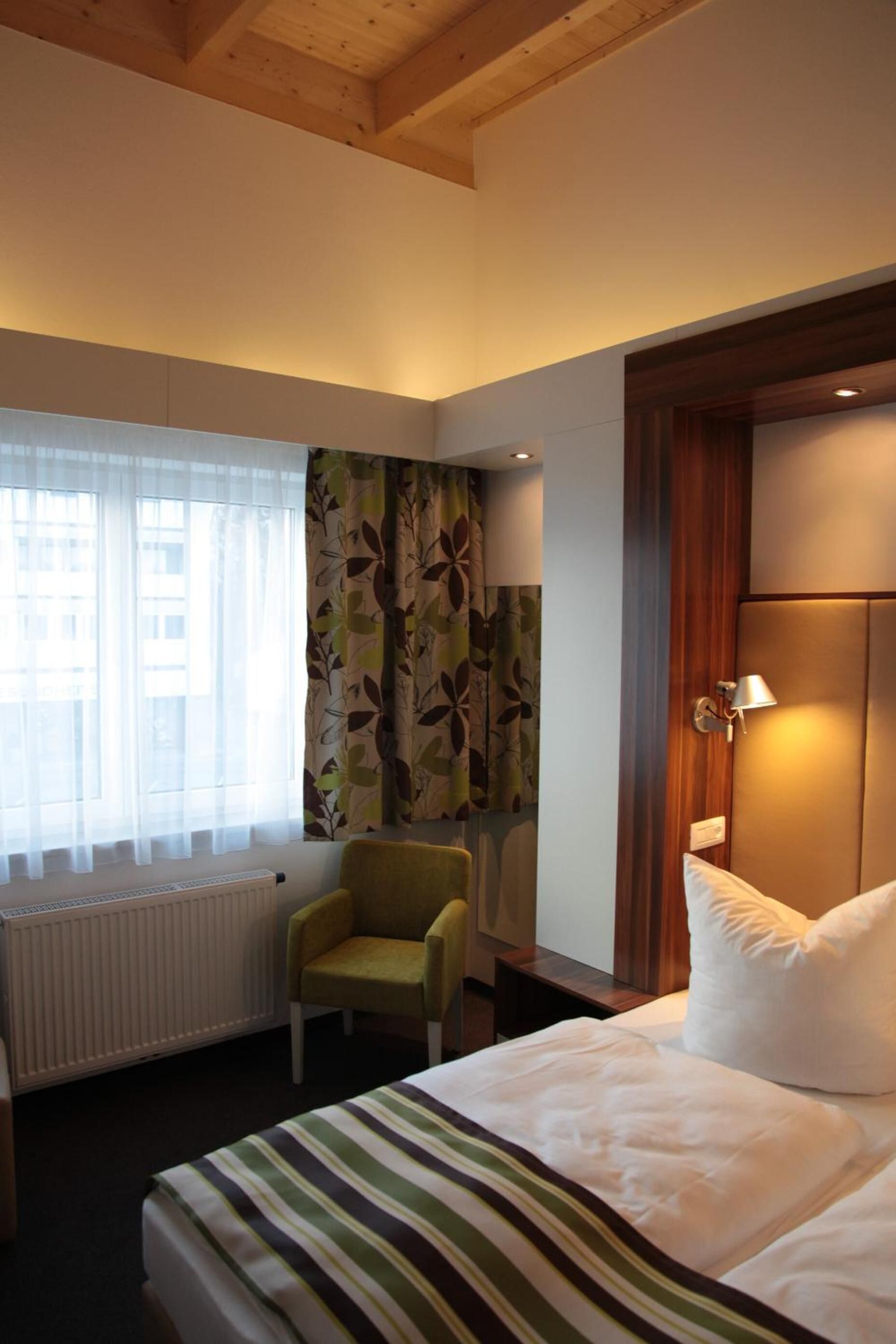 A hotel room with a bed, a chair, a radiator, and a window with curtains.