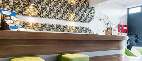 A reception area with a patterned wall, wooden reception desk, and green cushioned stools.