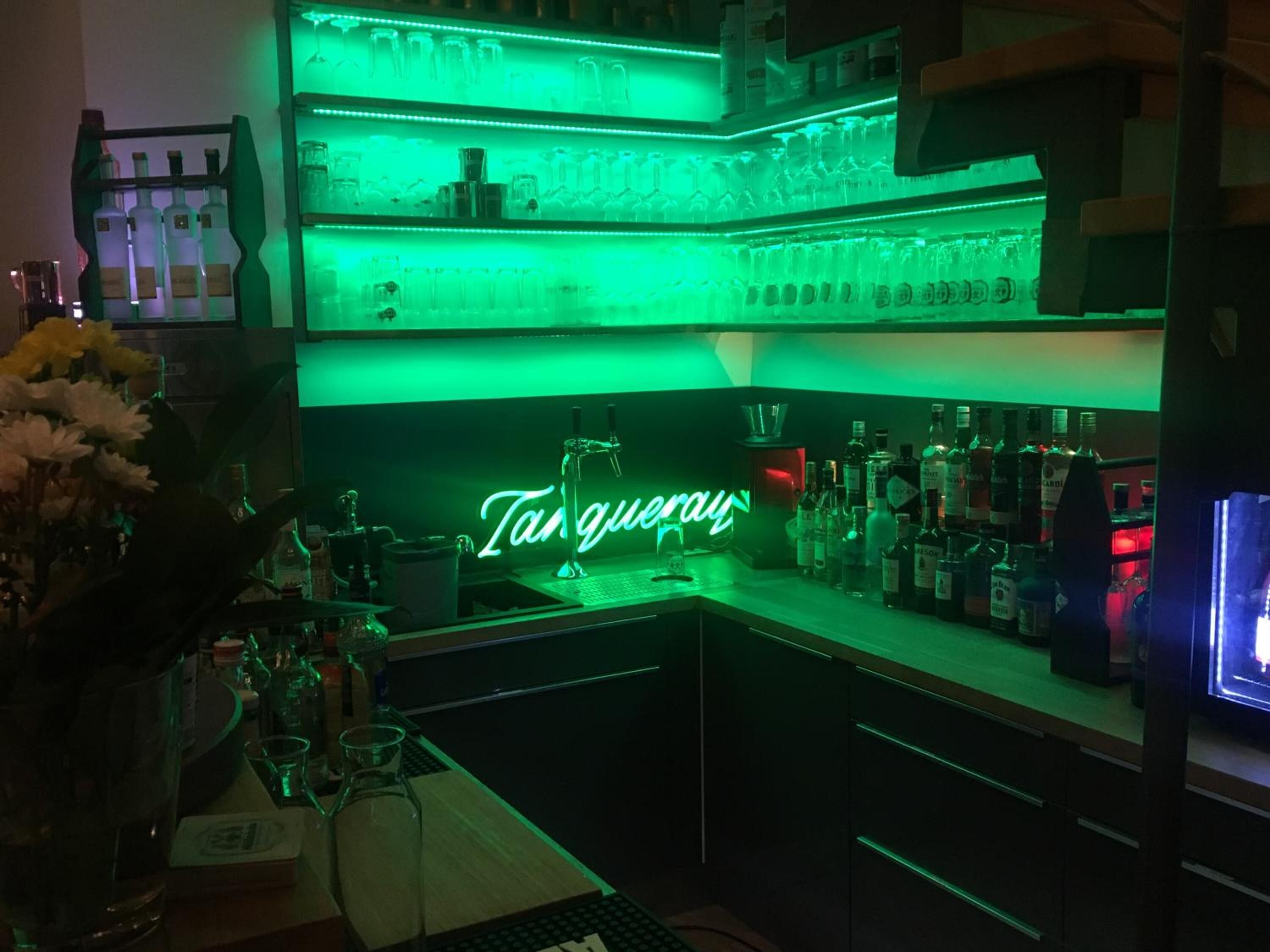 A bar area with shelves of bottles and glasses, a neon sign reading "Tanqueray", and a keyboard on the counter.