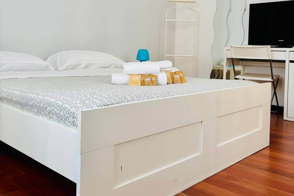 A neatly made bed with white bedding and a patterned cover, a bedside table with a blue lamp, and a desk with a flat-screen TV.