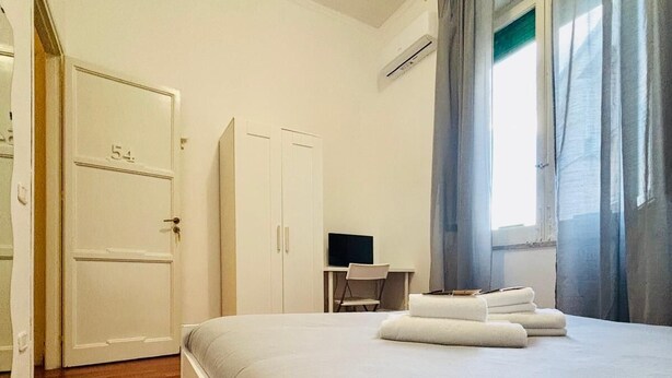A hotel room with a bed, a desk, a chair, a wardrobe, and a window with curtains. - Castelnuovo Rooms  (Palermo)