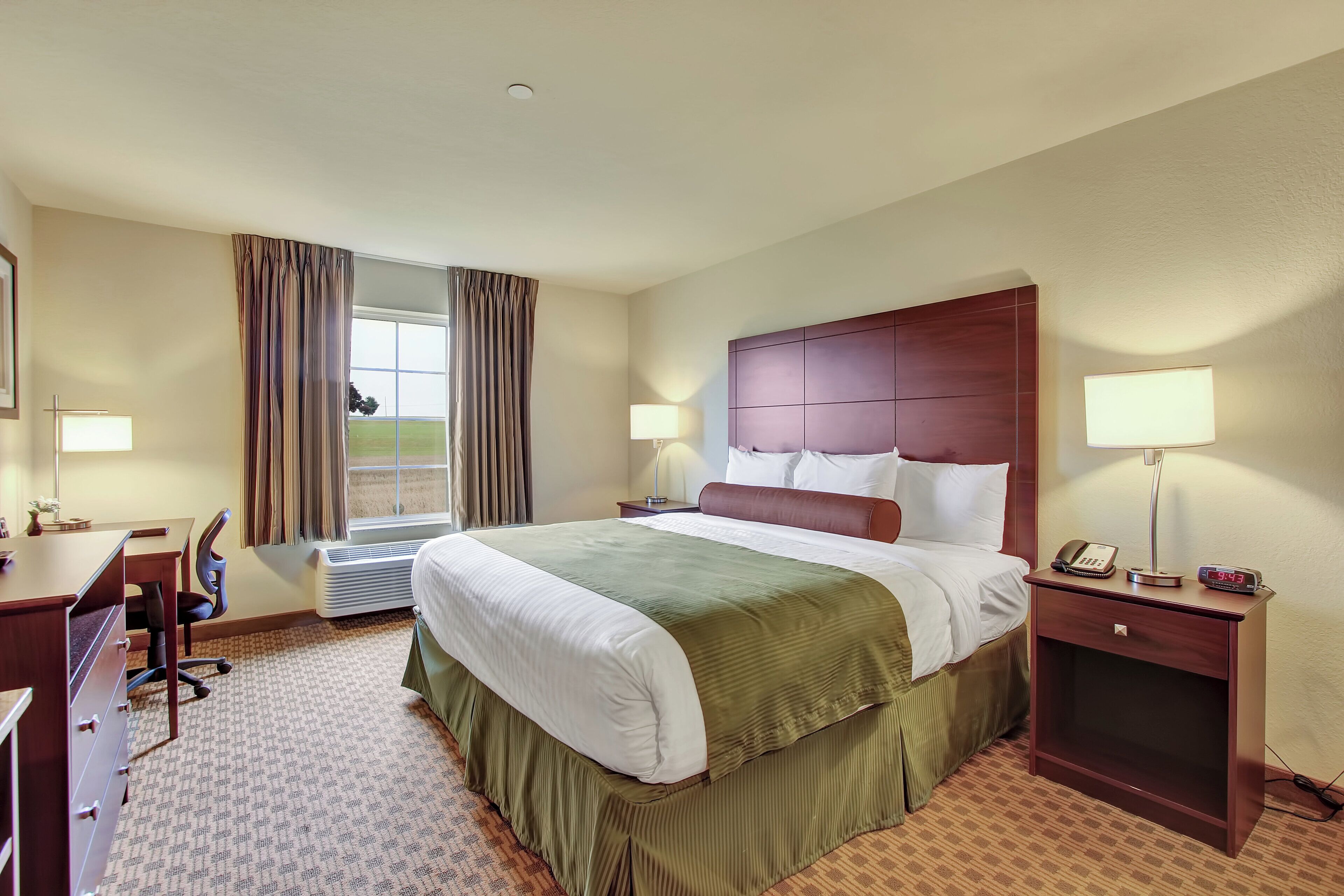Ein Hotelzimmer mit einem großen Bett, einem Schreibtisch, einem Sessel, einem Nachttisch mit Lampe und einem Fenster mit Vorhängen.