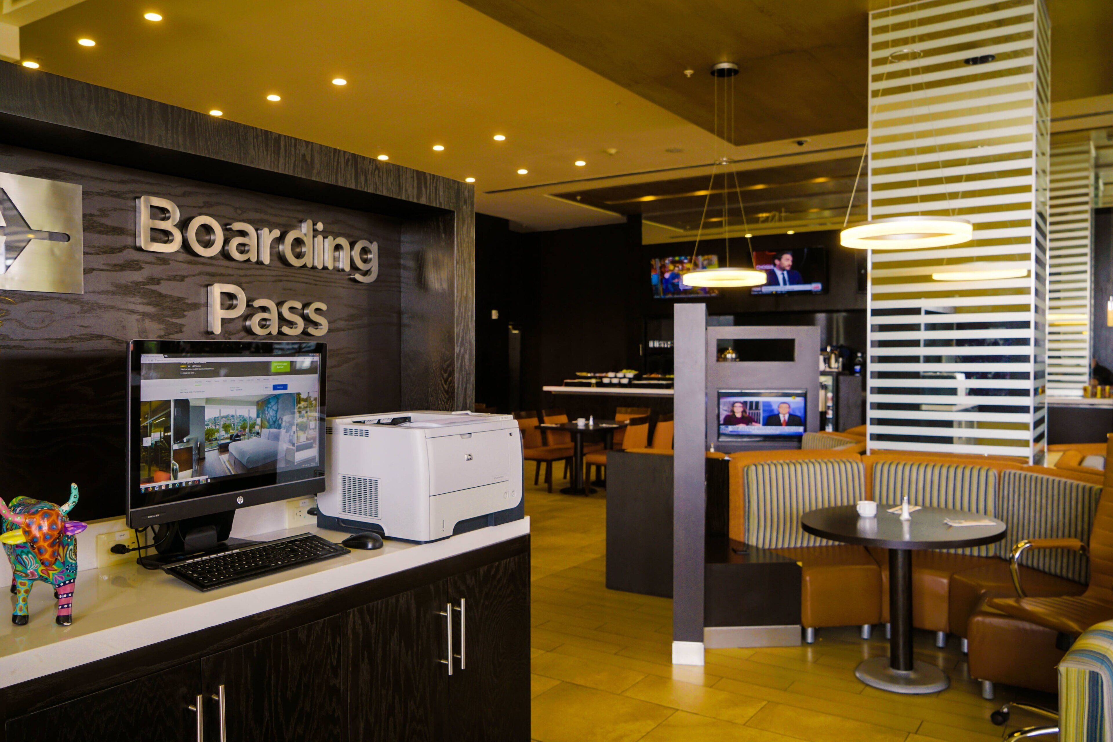 Un moderno vestíbulo de hotel con un letrero que dice 'Boarding Pass', un mostrador de recepción con computadora e impresora, áreas de estar y un televisor mostrando noticias.