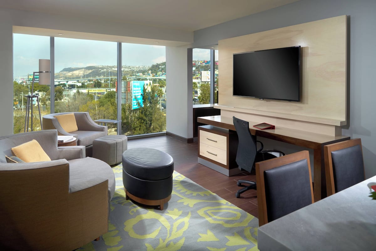 A modern hotel room with a large TV, comfortable seating, a desk, and a view of the city.