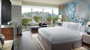 A modern hotel room with a large bed, a flat-screen TV, a sofa, and a city view through large windows. - Courtyard by Marriott Queretaro (Querétaro)