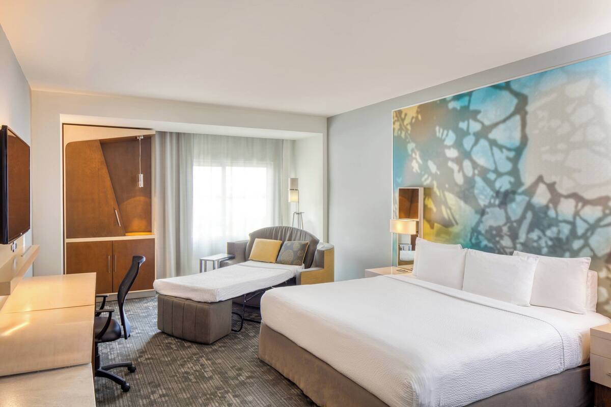A hotel room with a large bed, a desk, a chair, and a wardrobe.