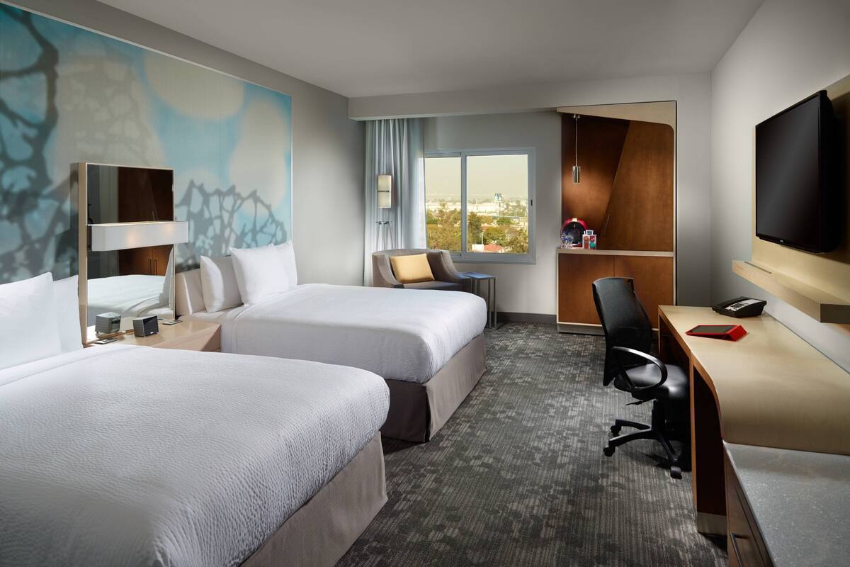 A hotel room with two beds, a desk, a TV, and a window with a city view.
