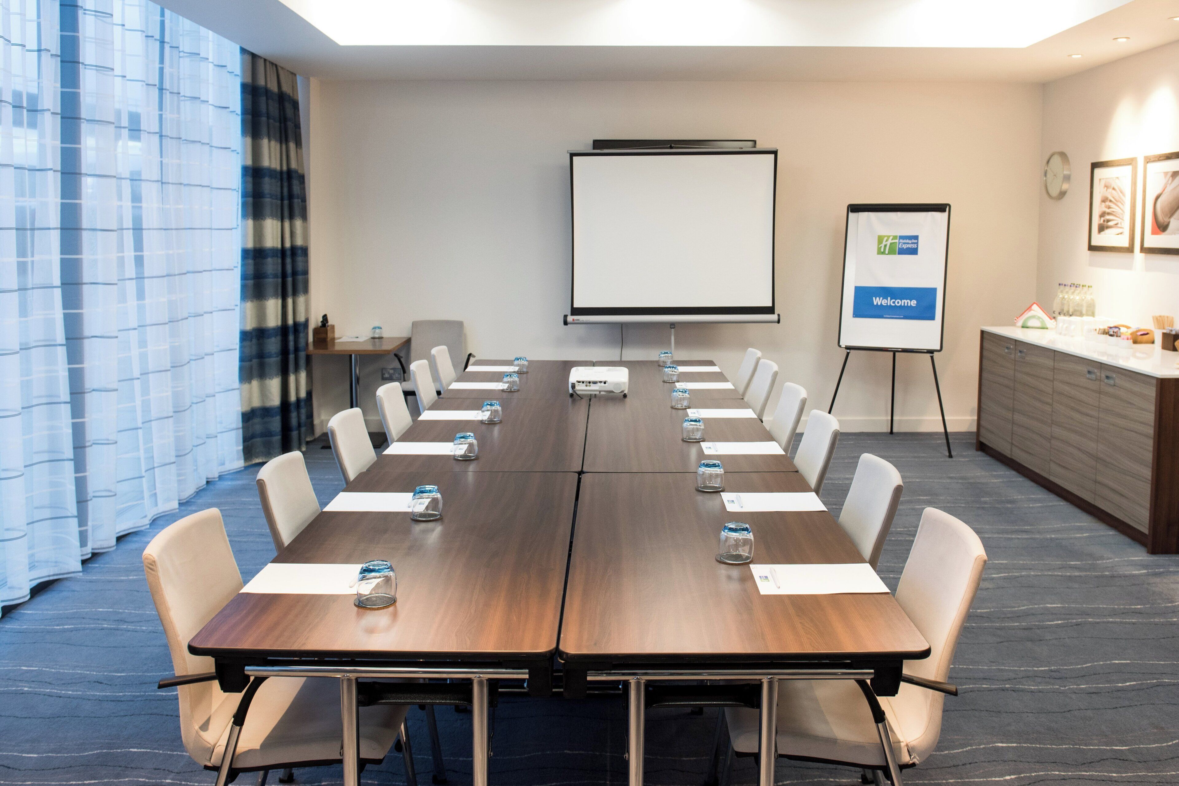 A conference room with a long table, chairs, a projector screen, and a welcome sign.