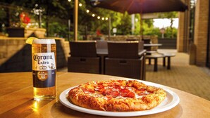 Une bouteille de bière Corona Extra et une pizza aux pepperoni sur une table dans un espace extérieur aménagé avec des guirlandes lumineuses et des parasols.