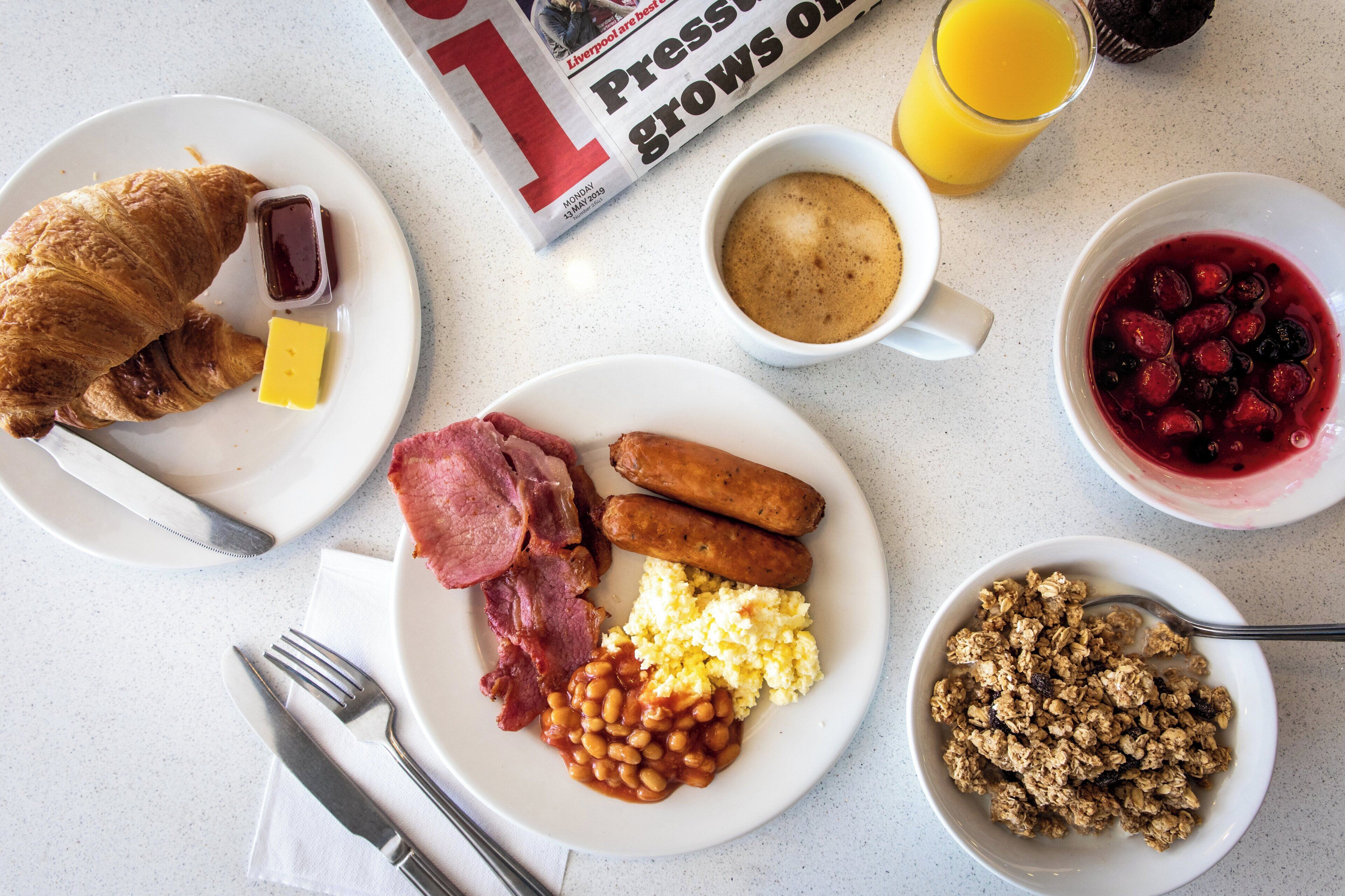 Une table de petit-déjeuner avec des croissants, du jambon, des saucisses, des haricots, des œufs brouillés, du jus d’orange, du café et un bol de granola.