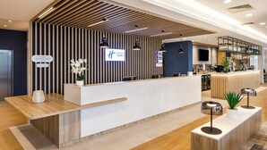 A modern hotel reception area with a wooden reception desk, a digital sign displaying the hotel name, and a bar area in the background. - Holiday Inn Express London - ExCeL by IHG (London)