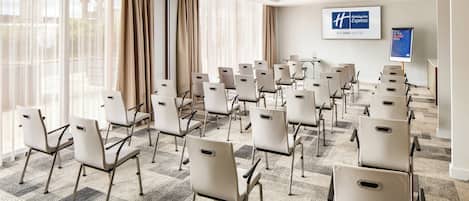 A conference room with rows of chairs, large windows with curtains, and a Holiday Inn Express sign on the wall.