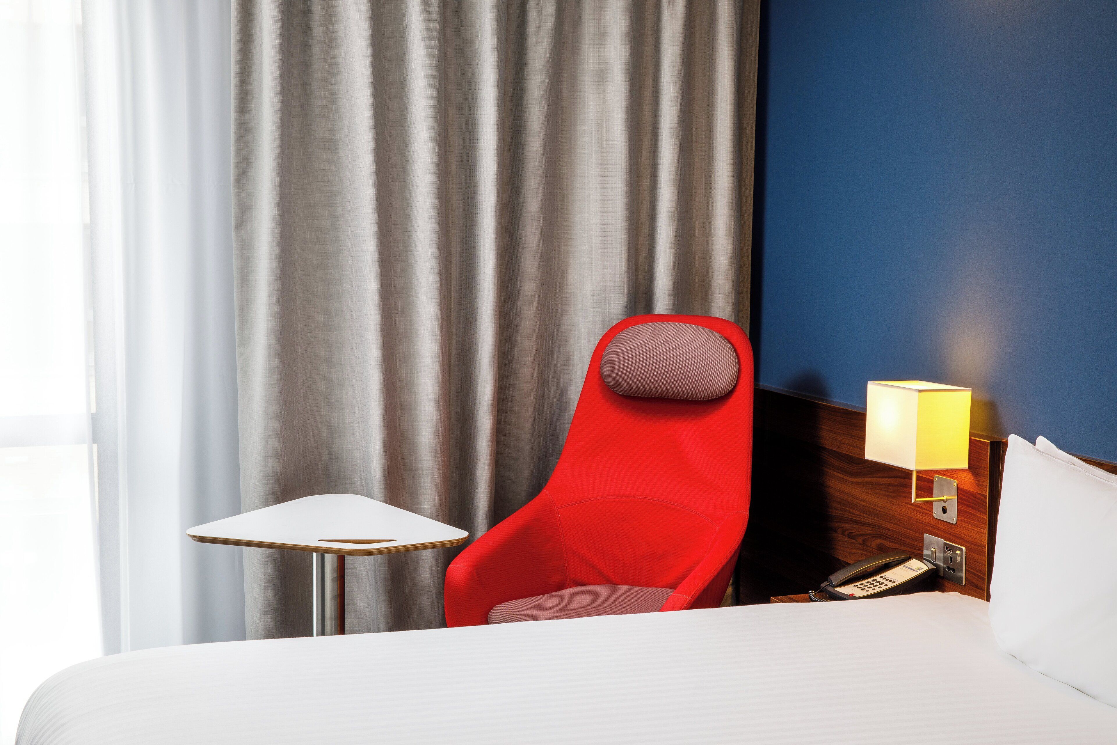 A hotel room with a bed, a red chair, a small table, a lamp, and a telephone.