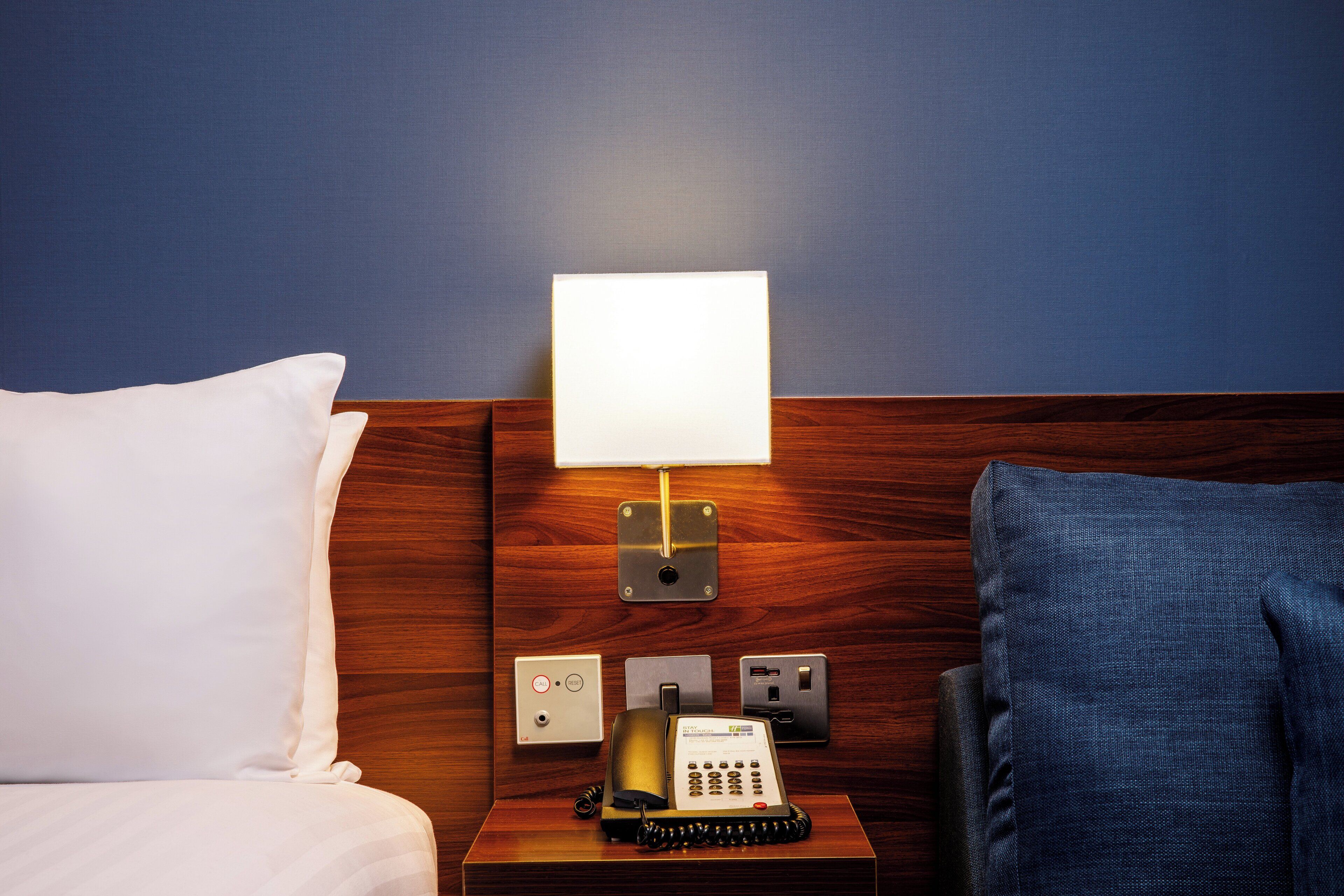 Habitación de hotel con cabecera de madera, mesita de noche con lámpara, teléfono y toma de corriente.