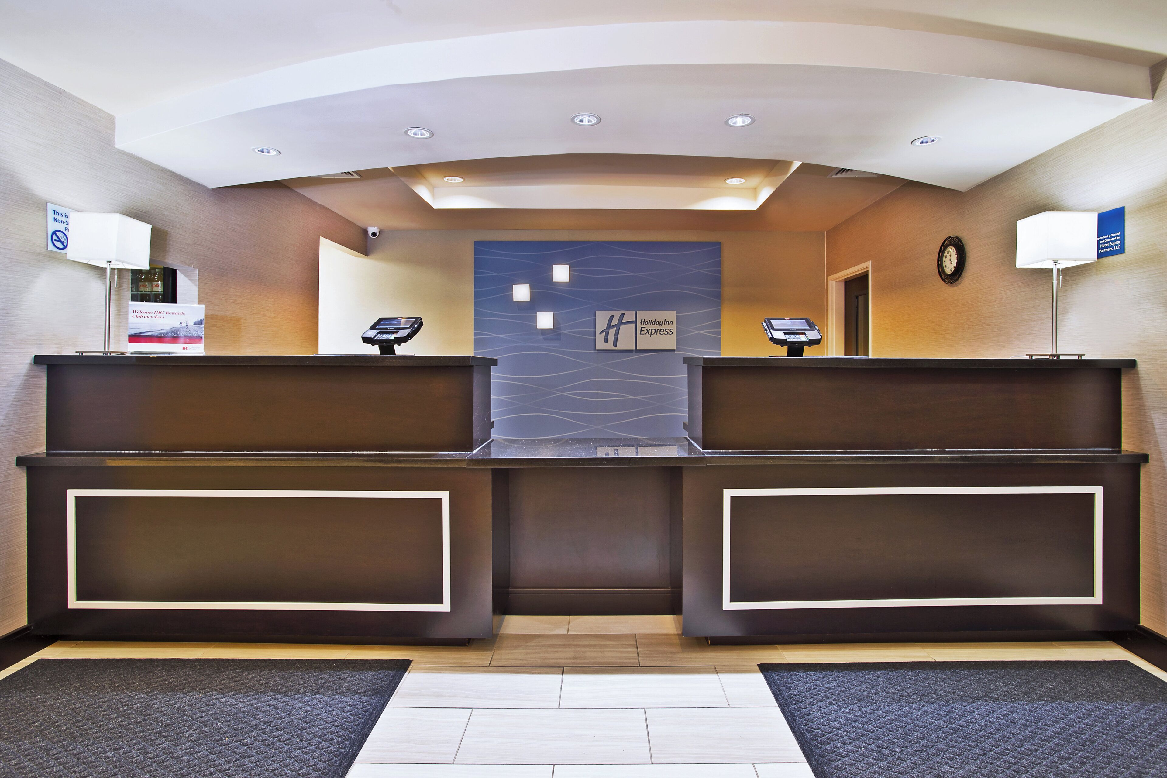 A reception area with a modern design, featuring a central desk with a digital display, two side desks, a clock on the wall, and a sign indicating the hotel name.