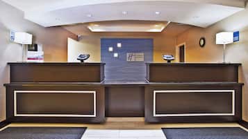 A reception area with a modern design, featuring a central desk with a digital display, two side desks, a clock on the wall, and a sign indicating the hotel name.