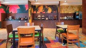 A hotel dining area with wooden tables and chairs, a counter with various food items, and a fruit-themed wall mural. - Fairfield Inn & Suites Fredericksburg (Fredericksburg)