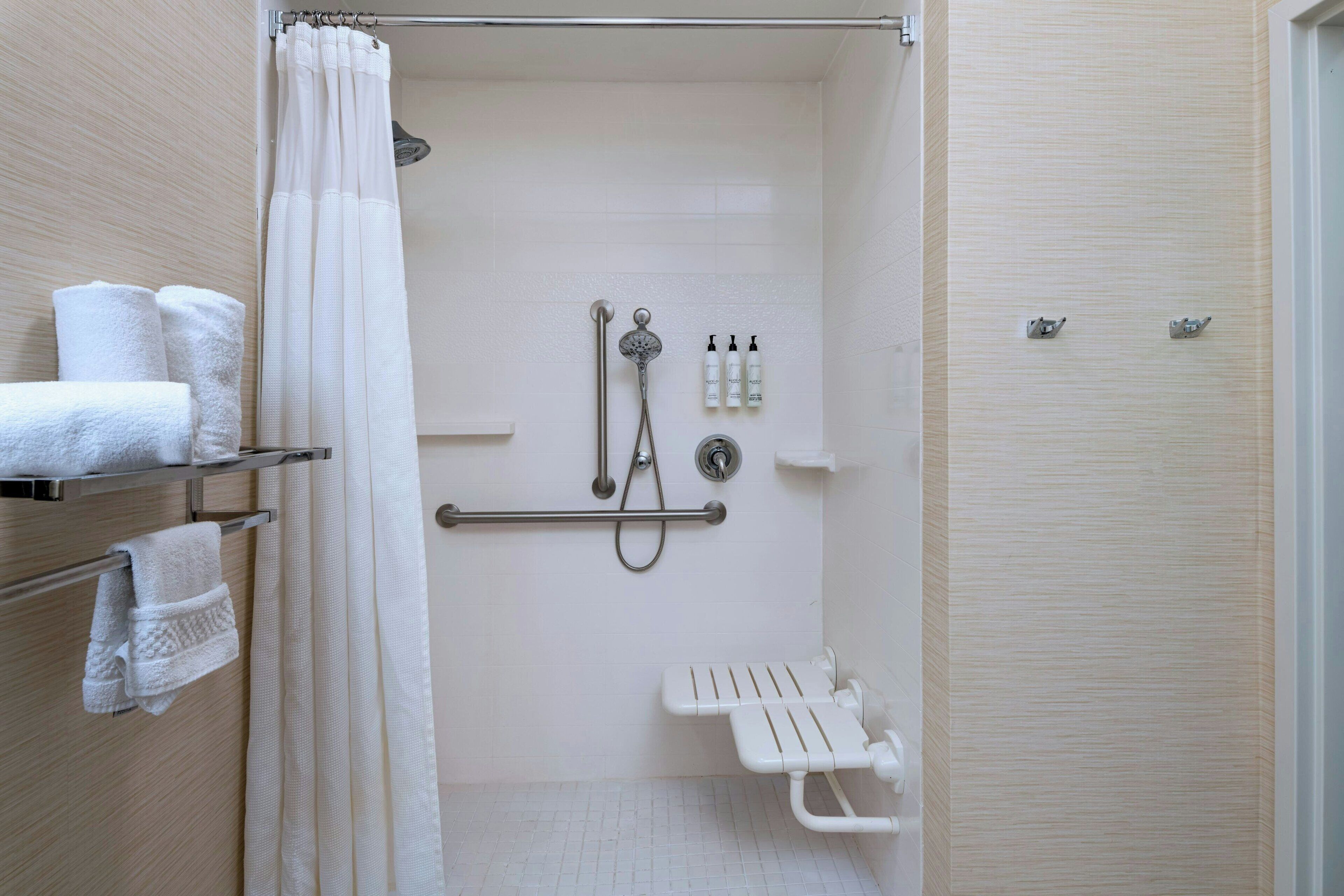 Une douche à l’italienne est équipée d’un pommeau de douche, d’un pommeau de douche à main et d’un flexible de douche. Trois bouteilles de shampoing et d’après-shampoing sont posées sur le mur. Un banc blanc est placé dans l’espace de la douche.