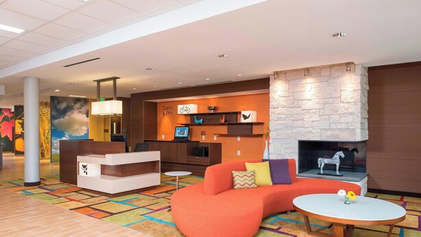 A modern lobby with a curved orange sofa, a round wooden table, and a stone fireplace. - Fairfield Inn & Suites Fredericksburg (Fredericksburg)