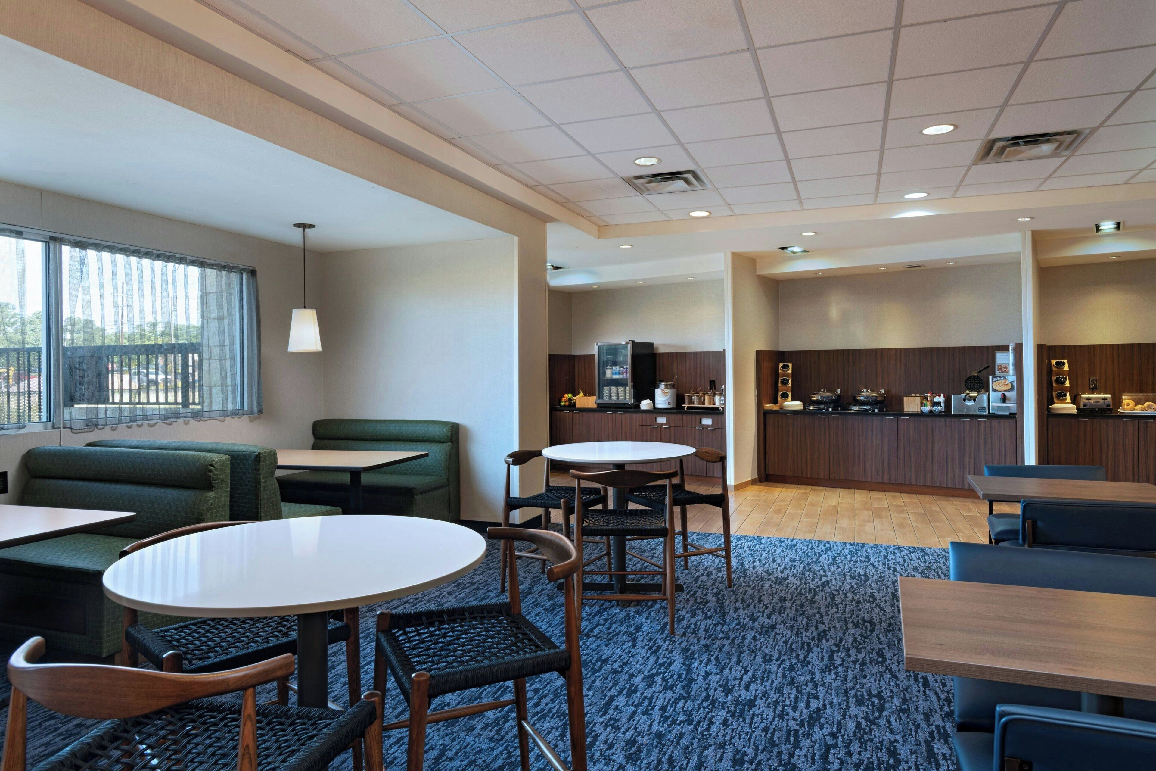 A hotel lobby with seating areas, a kitchen in the background, and a large window with a view.