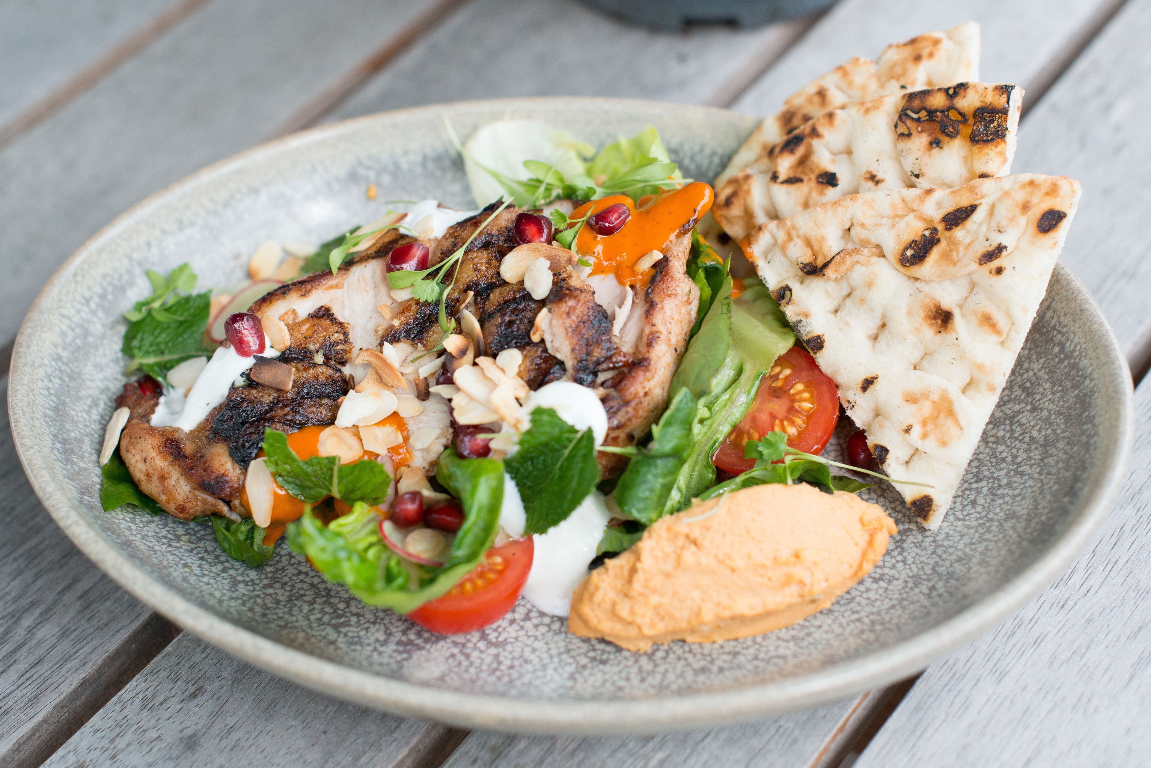 A plate of grilled chicken, fresh salad, pomegranate seeds, and pita bread on a wooden table.