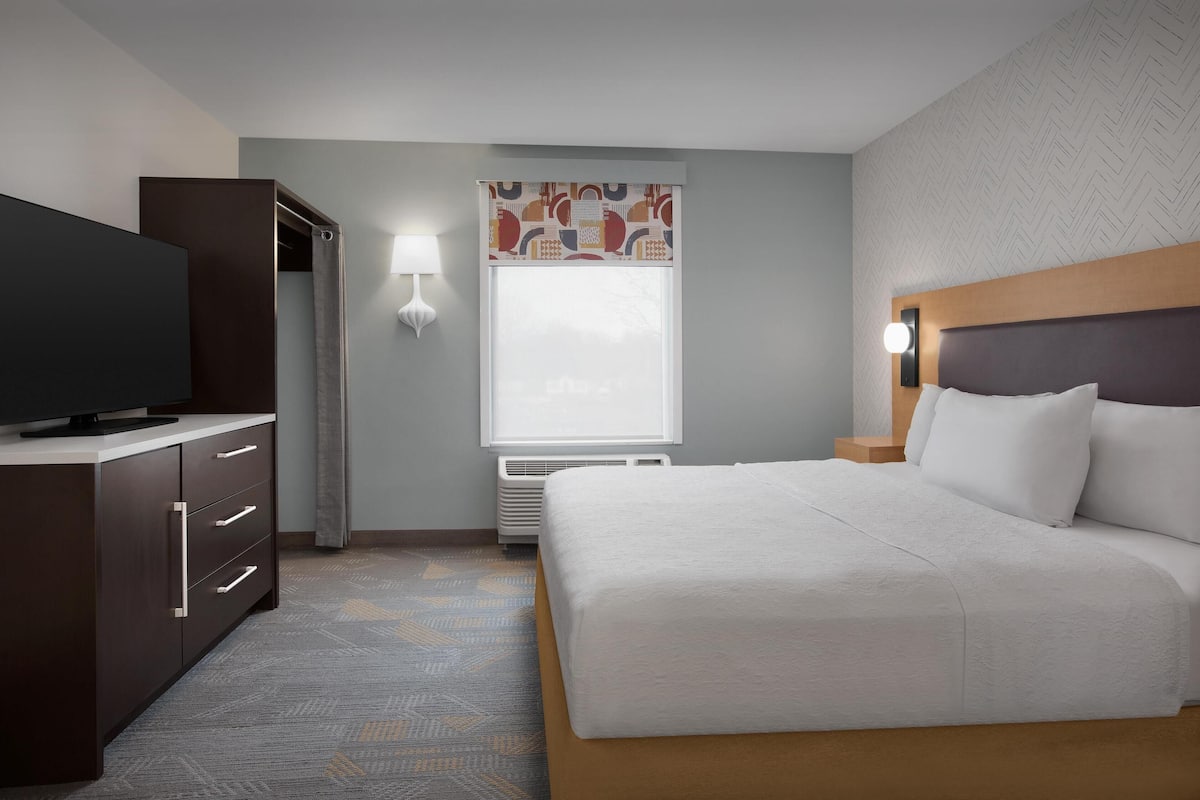A hotel room with a bed, a TV, a wardrobe, a window with a view, and a wall-mounted lamp.