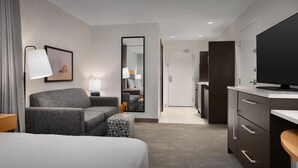 A modern hotel room with a bed, a sofa, a small table, a TV, and a bathroom visible through an open door.