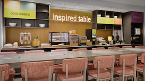 A breakfast area with a long table, orange chairs, and various breakfast items displayed on shelves.