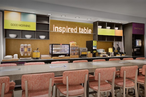 Un espace dédié au petit-déjeuner, avec une longue table, des chaises orange et divers aliments pour le petit-déjeuner présentés sur des étagères.