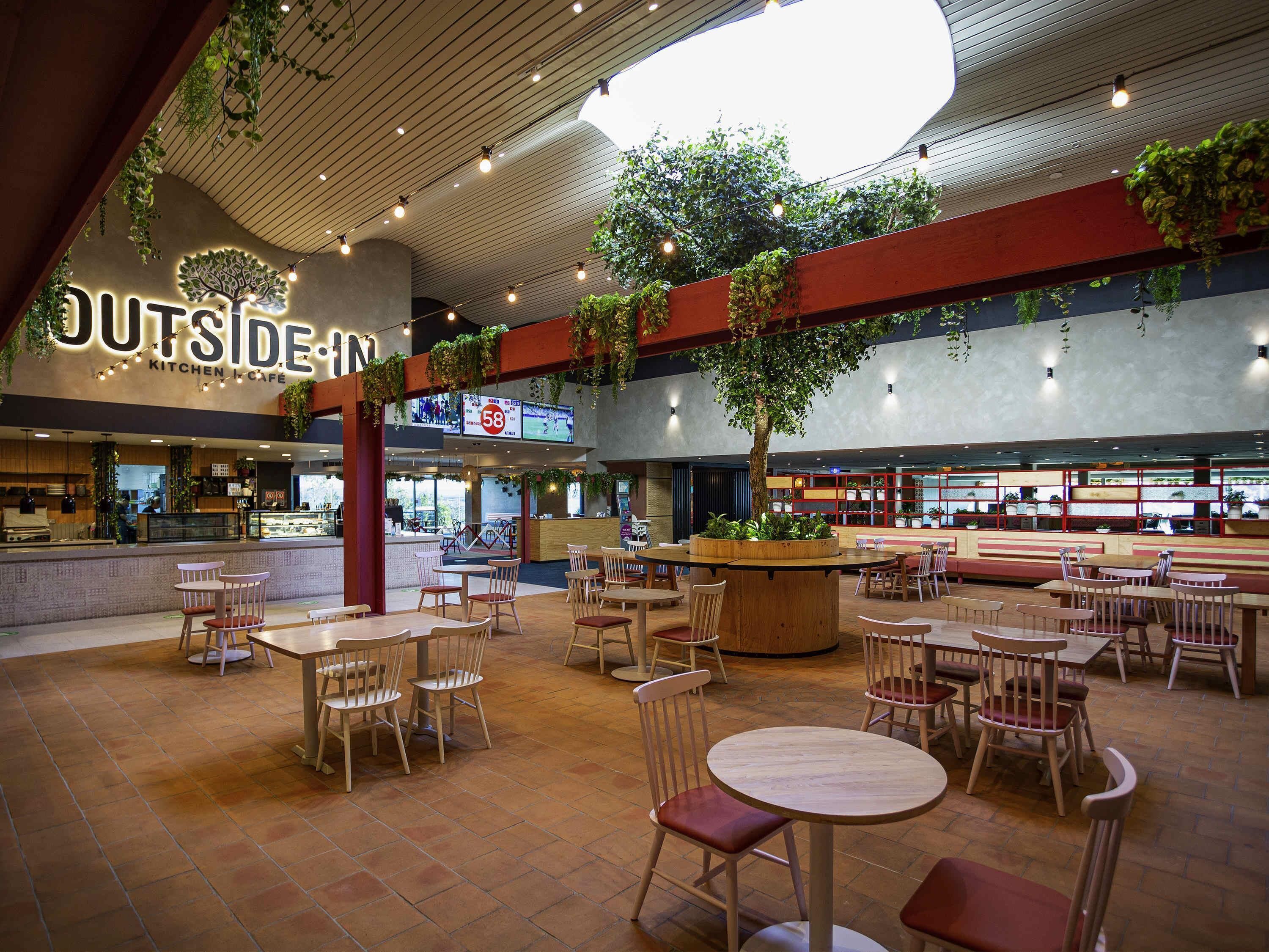 Un vaste espace de restauration intérieur, doté de nombreuses tables et chaises, d’un grand bac à plantes central avec un arbre, et d’un restaurant bien visible appelé « OUTSIDE IN Kitchen & Cafe ».