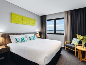 Habitación de hotel con una cama grande, una mesita de noche, un ventanal con cortinas, vista al mar y una pequeña mesa redonda con un jarrón de flores.