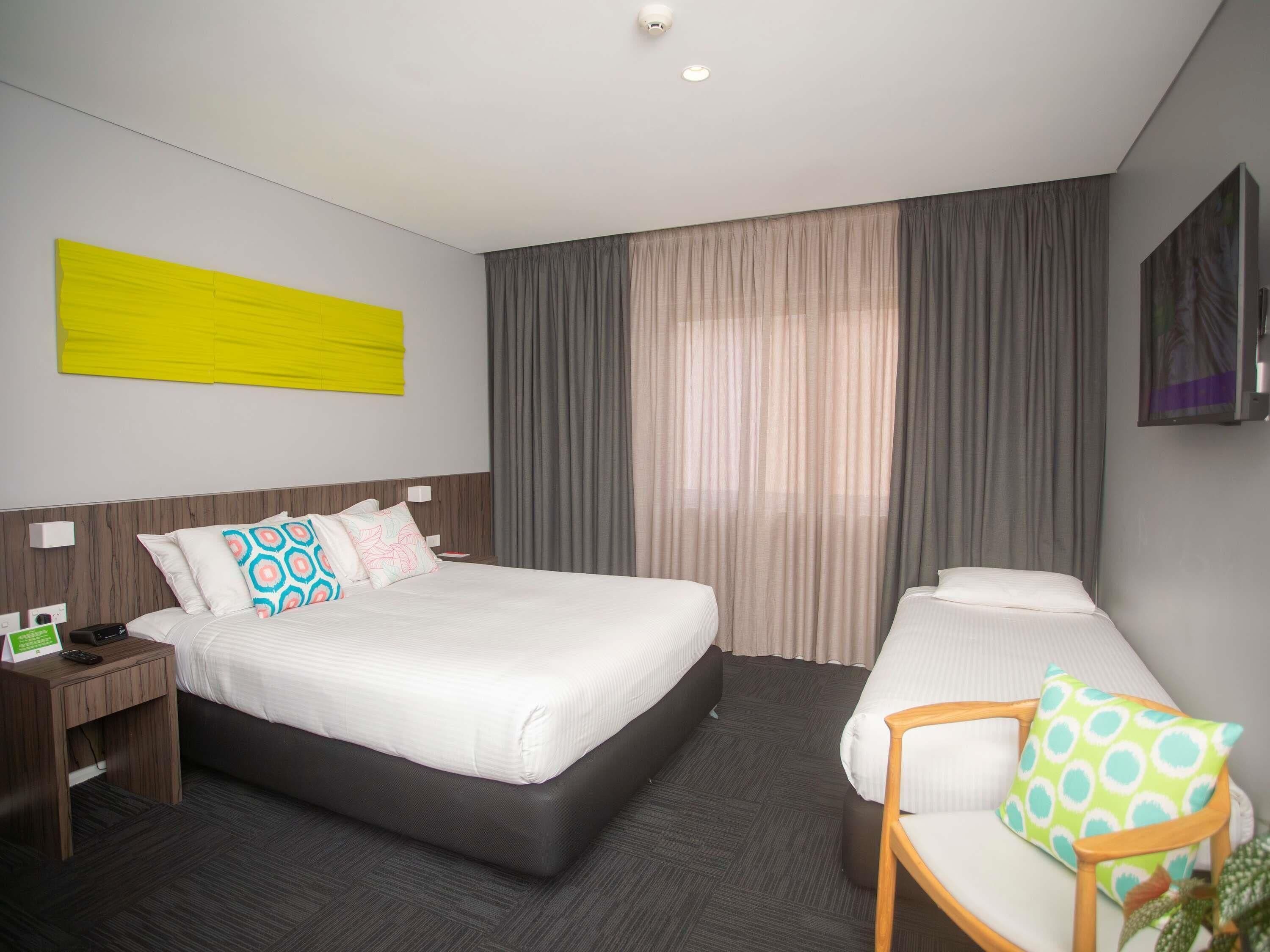 Habitación de hotel con una cama grande, una mesita de noche con un teléfono y un cartel, una silla con cojín y un ventanal con cortinas.