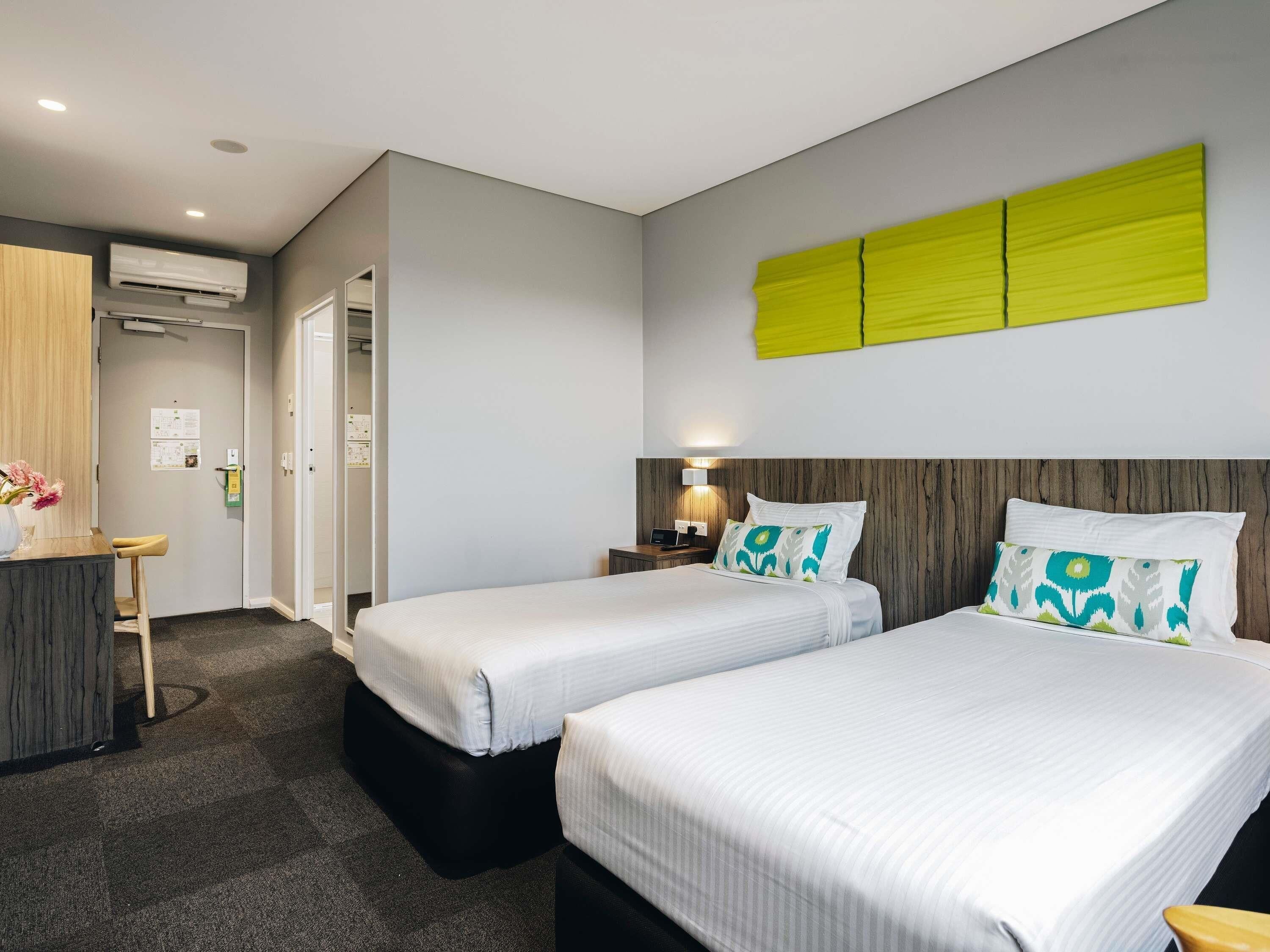 Une chambre d’hôtel avec deux lits, un bureau, une chaise, un appareil de climatisation et un luminaire fixé au mur.