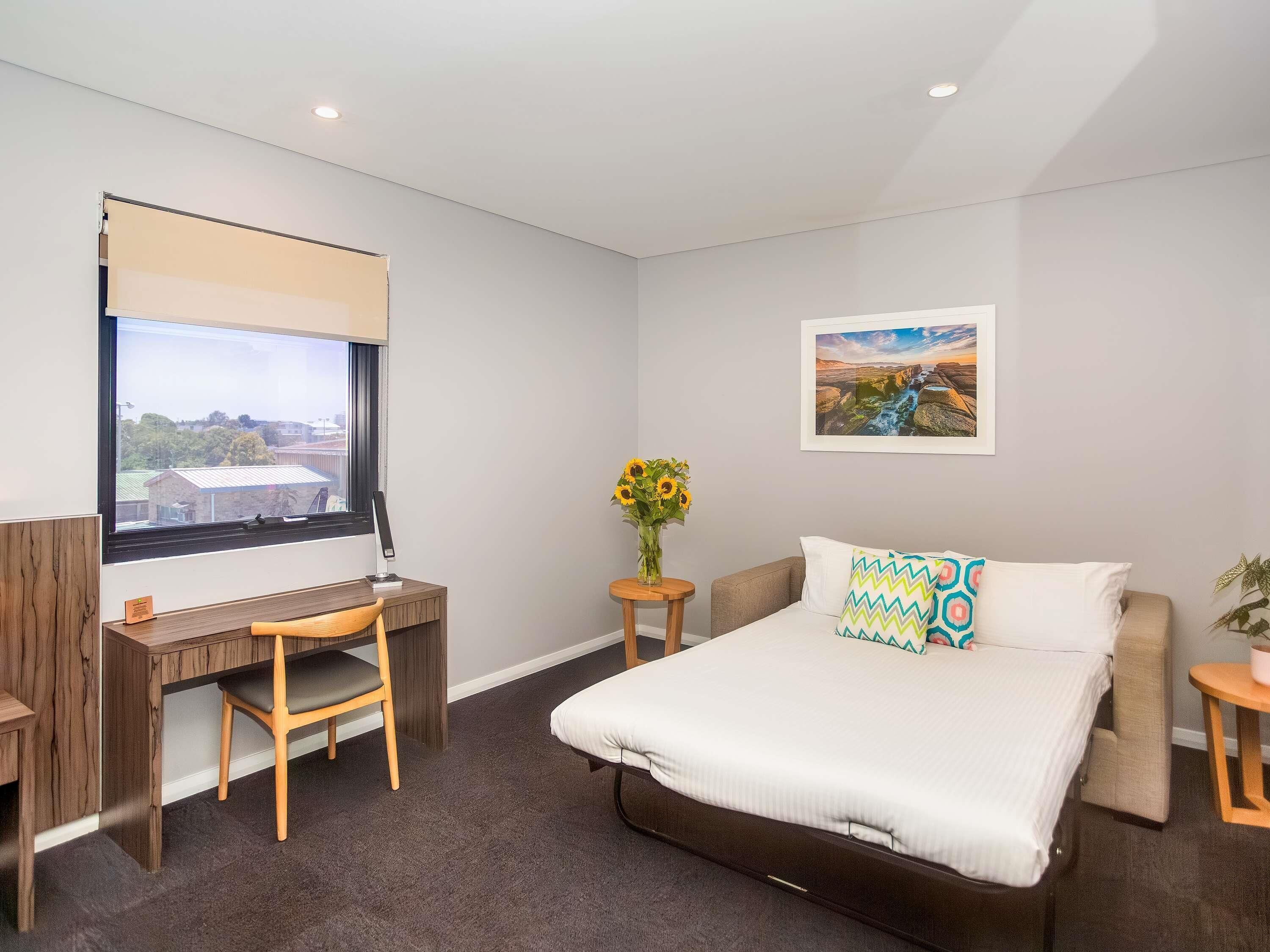 A hotel room with a bed, a desk, a chair, a window with a view, a framed picture on the wall, and a vase with sunflowers.