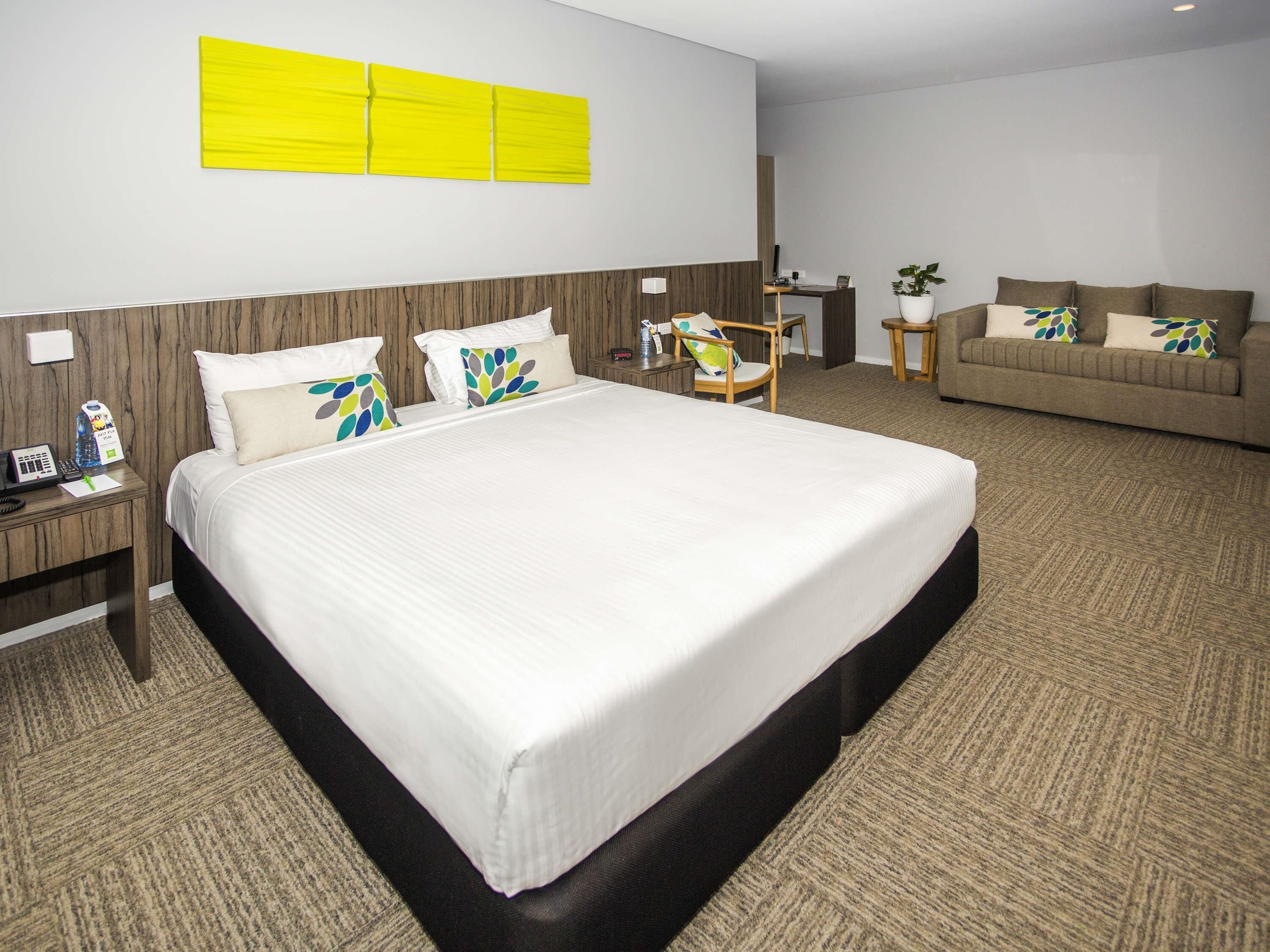 A hotel room with a large bed, a wooden headboard, a desk, a chair, a sofa, and a potted plant.