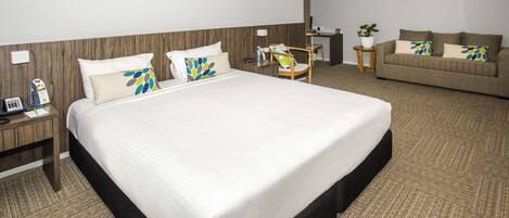A hotel room with a large bed, a wooden headboard, a desk, a chair, a sofa, and a potted plant.