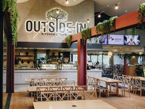 A modern cafe with a prominent sign reading "OUTSIDE IN KITCHEN CAFÉ", featuring a tree logo, wooden furniture, and indoor plants. - ibis Styles The Entrance (Long Jetty)
