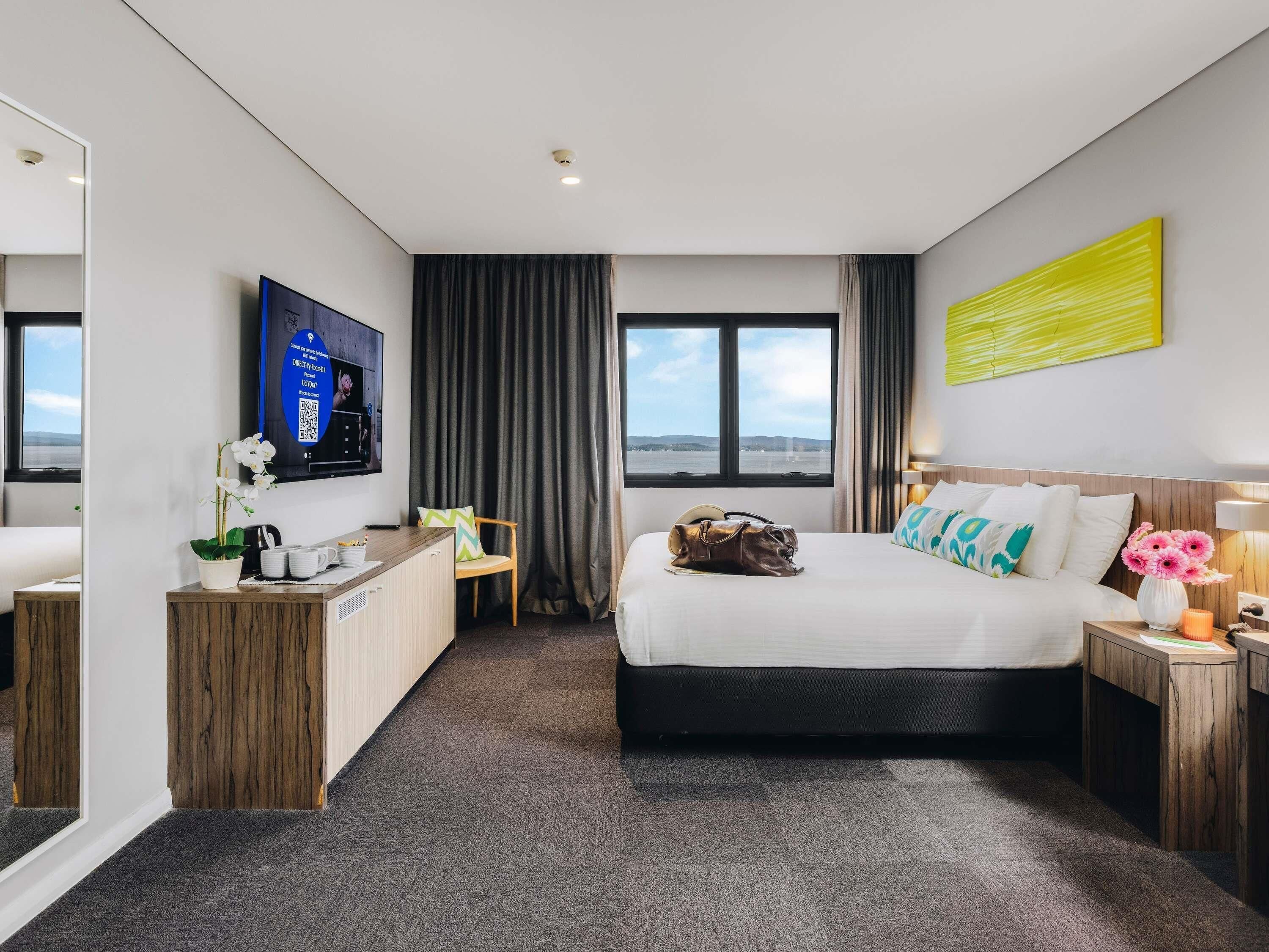 A hotel room with a bed, a desk, a TV, a window with a view, and a decorative wall piece.