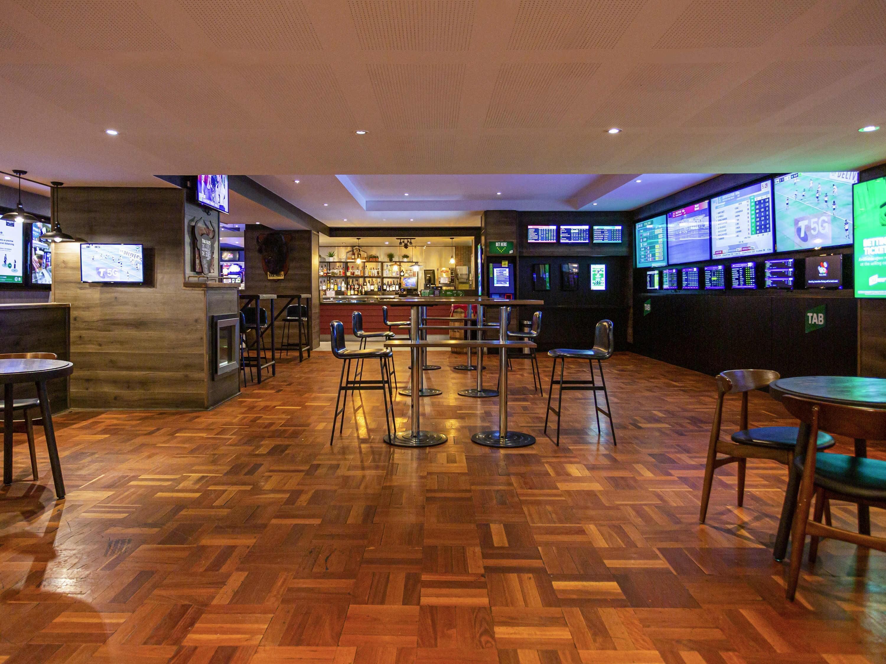 Zona de bar con suelo de madera, varios taburetes y una barra con un bartender.