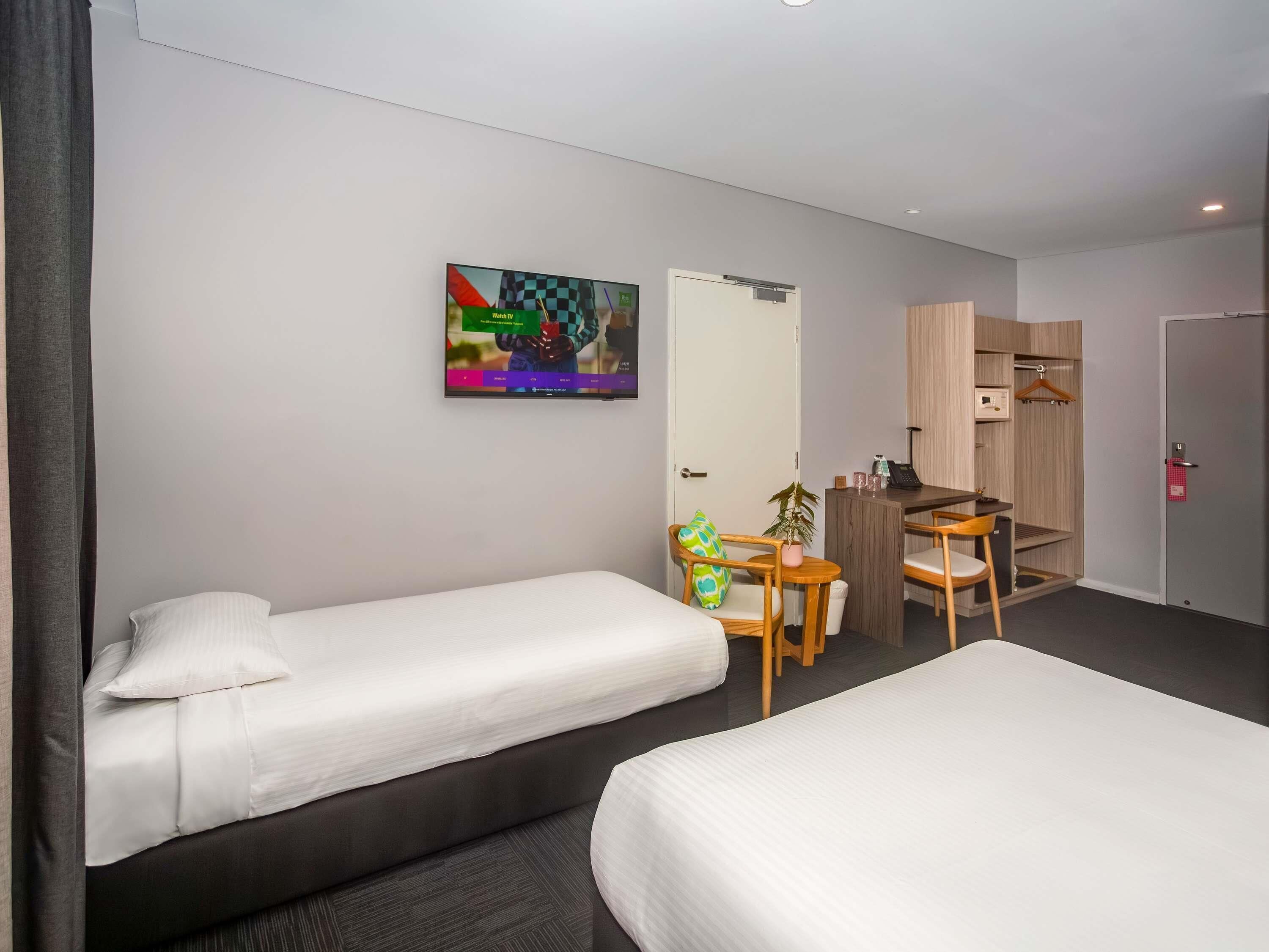 A hotel room with two single beds, a desk with a chair, a small table with a plant, a wardrobe, and a television on the wall.