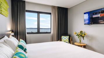 Habitación de hotel con cama, televisor montado en la pared, ventana con cortinas y una mesita con un jarrón de flores.