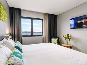 A hotel room with a bed, a TV mounted on the wall, a window with curtains, and a small table with a vase of flowers. - ibis Styles The Entrance (Long Jetty)