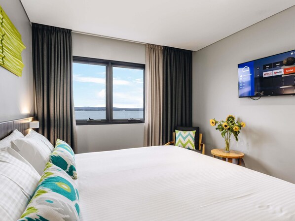 A hotel room with a bed, a TV mounted on the wall, a window with curtains, and a small table with a vase of flowers. - ibis Styles The Entrance (Long Jetty)