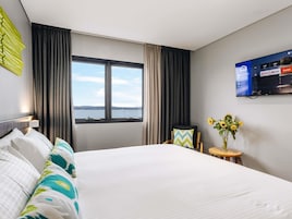 Habitación de hotel con cama, televisor montado en la pared, ventana con cortinas y una mesita con un jarrón de flores.