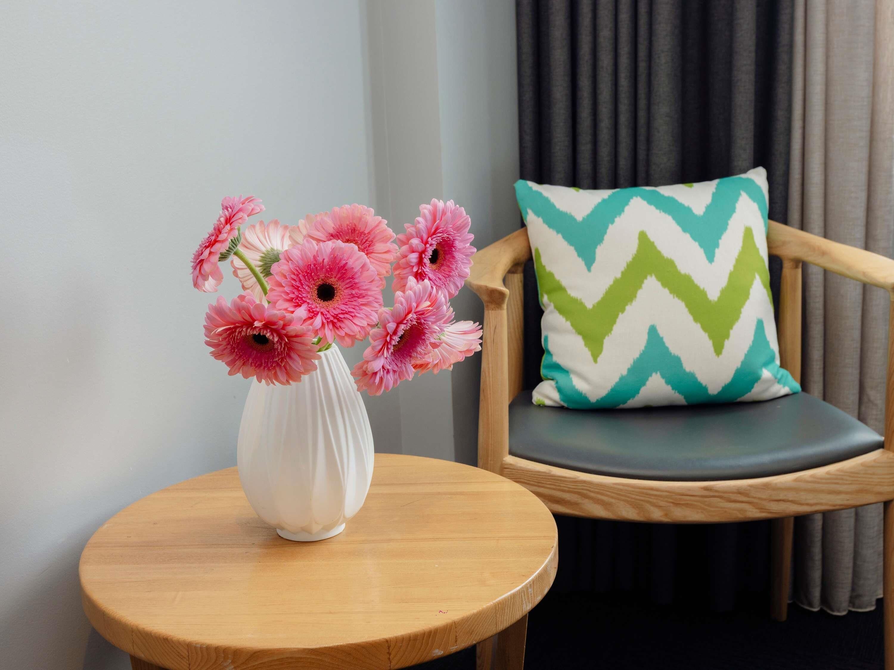 A round wooden table with a white vase containing pink flowers, a wooden chair with a green and white chevron pillow, and a grey curtain in the background.