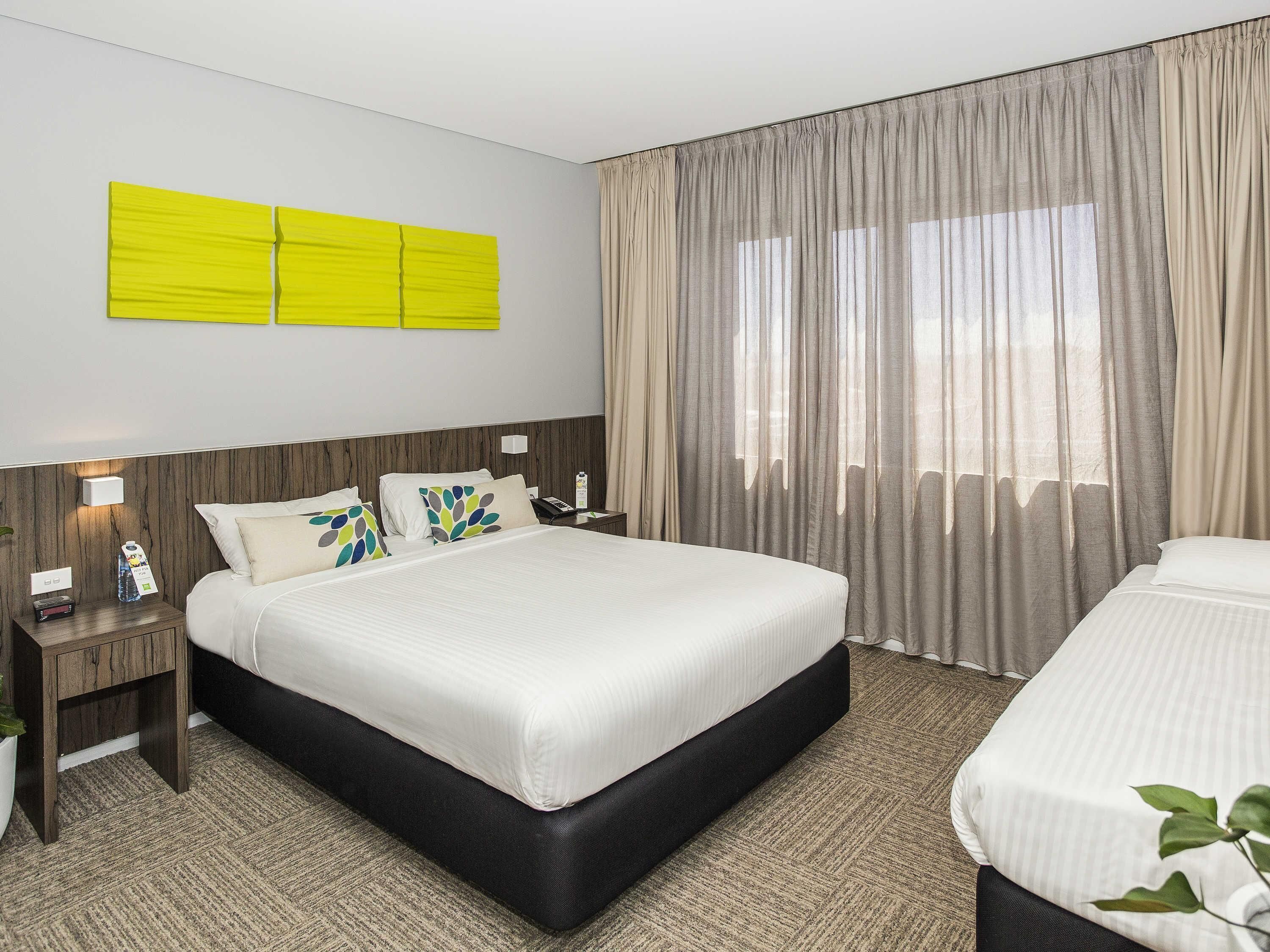 Une chambre d’hôtel avec un grand lit, des tables de chevet, une petite table, une lampe fixée au mur et une fenêtre avec des rideaux.