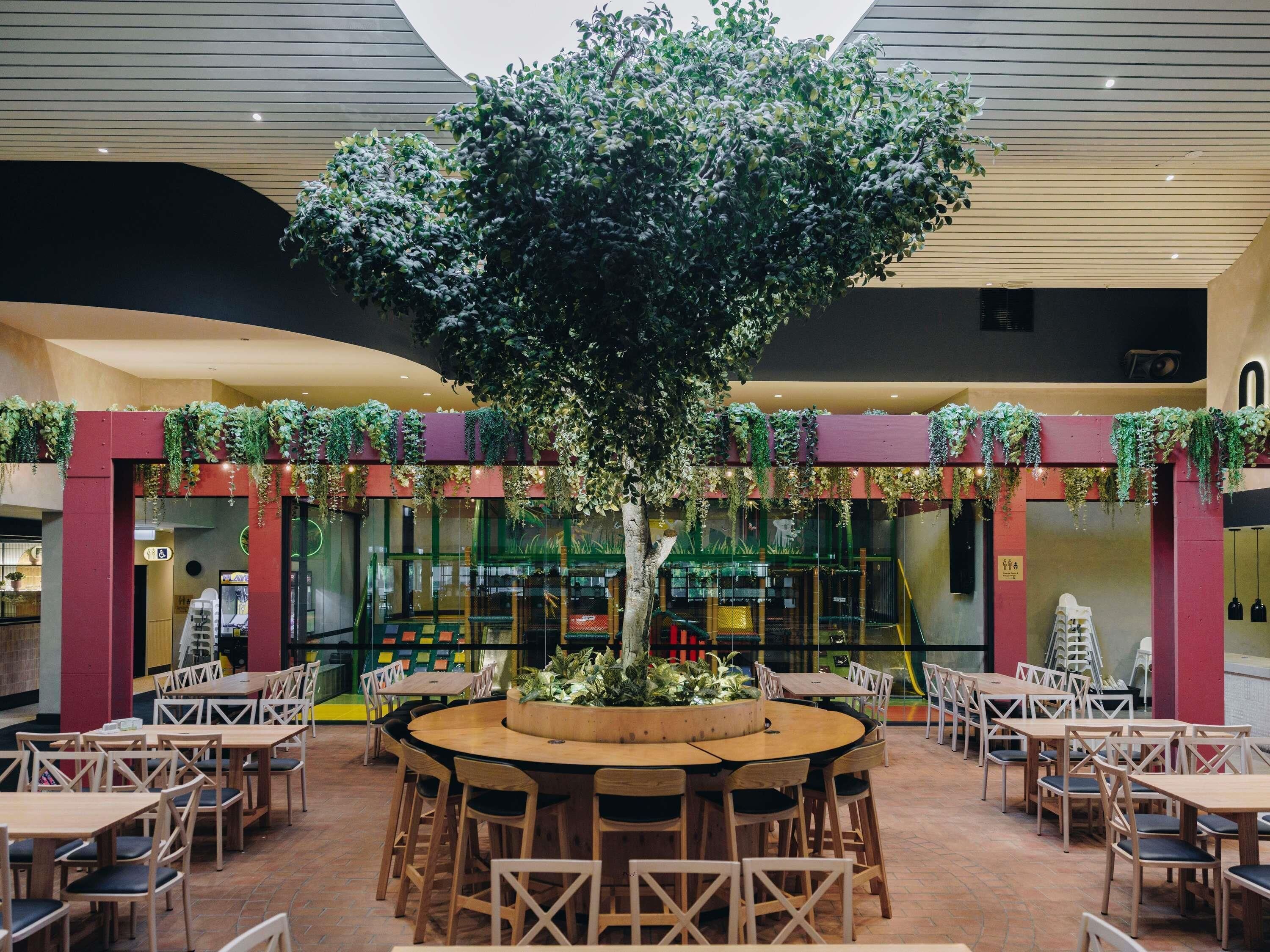 A restaurant with a central tree, surrounded by tables and chairs, and a glass wall in the background.