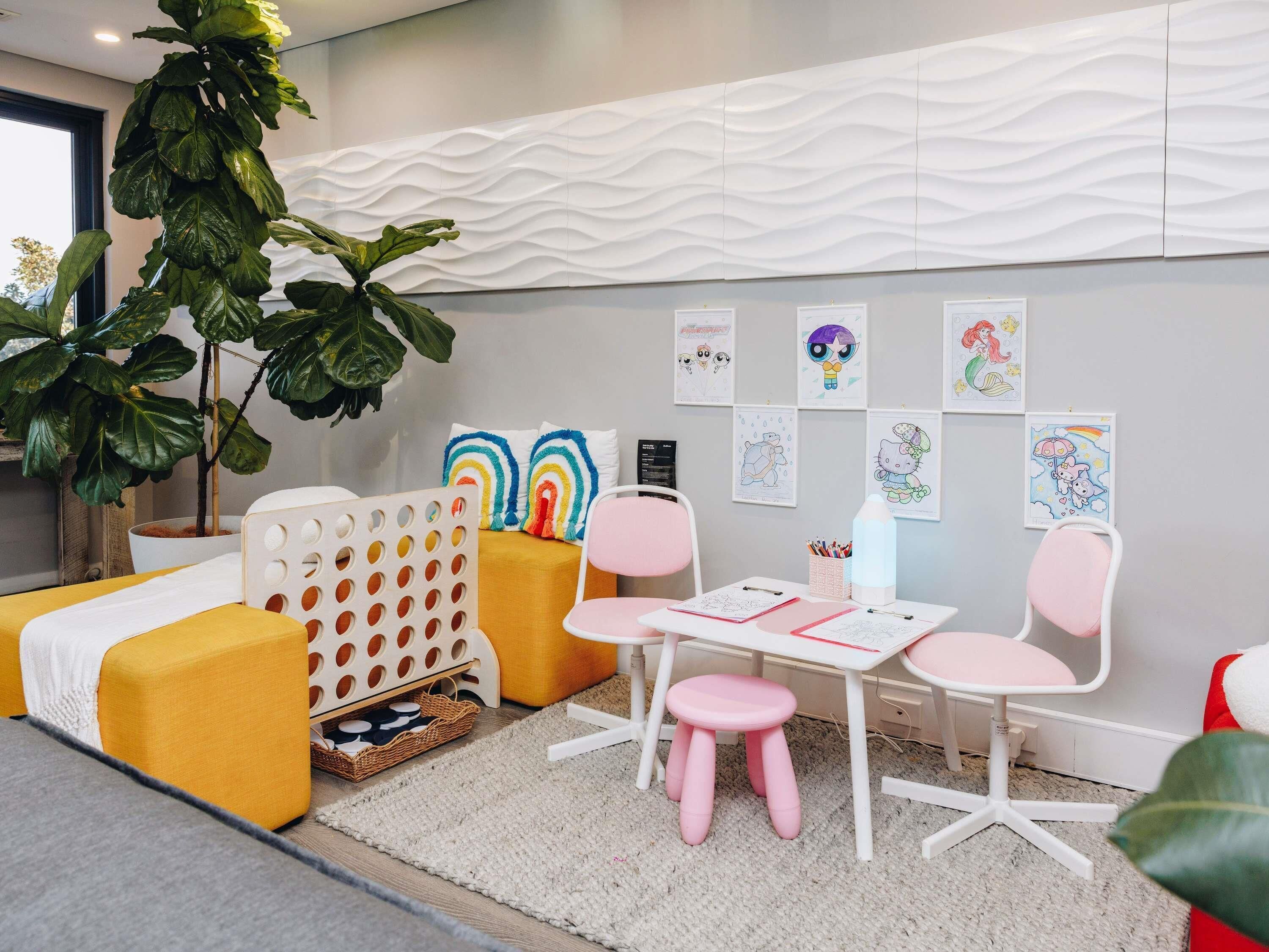 A children's playroom with a white wall, a large plant, a yellow couch, a white table with pink chairs, and various children's artwork.