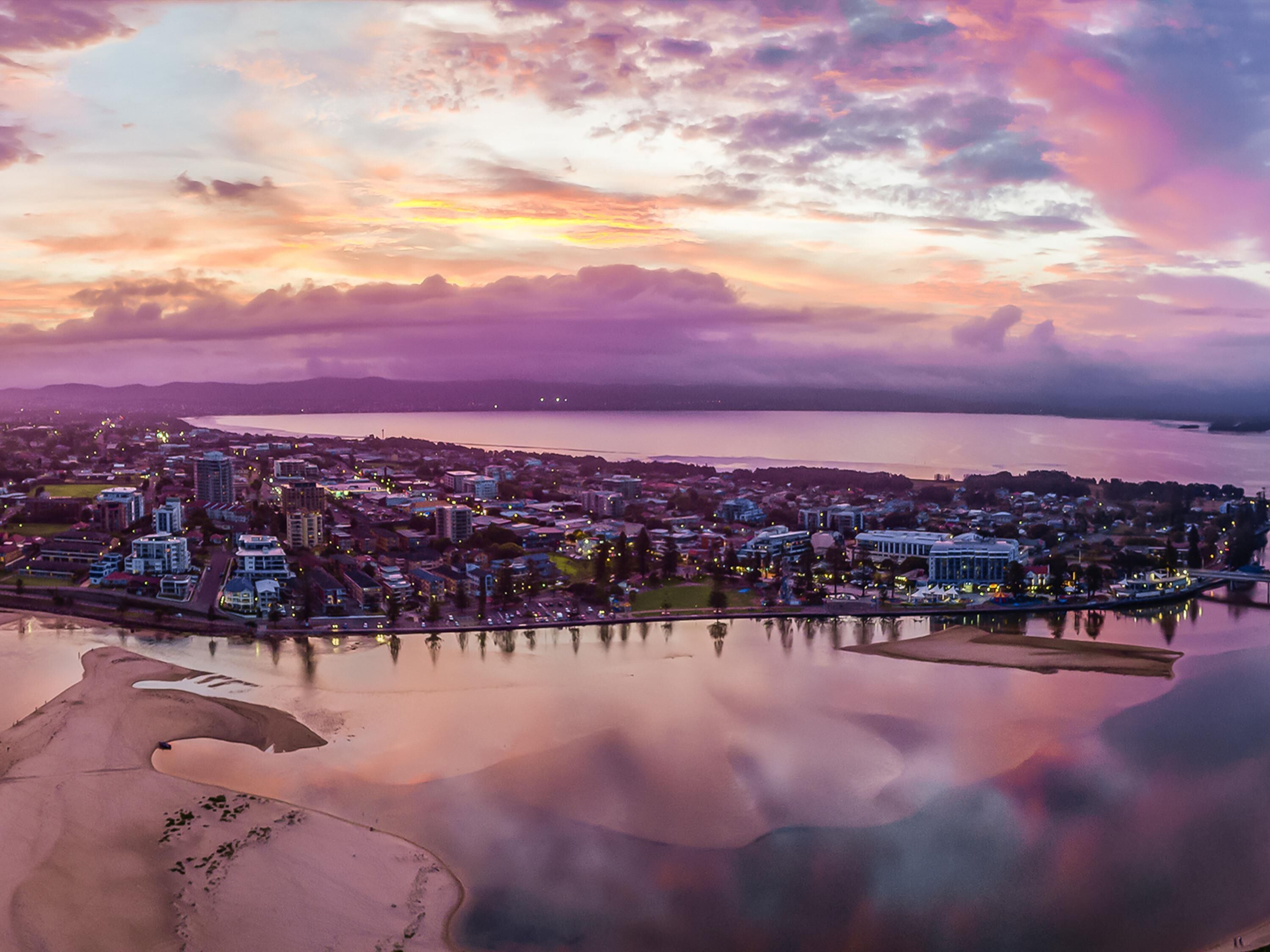 A coastal cityscape with a body of water reflecting the sky, buildings, and a bridge.