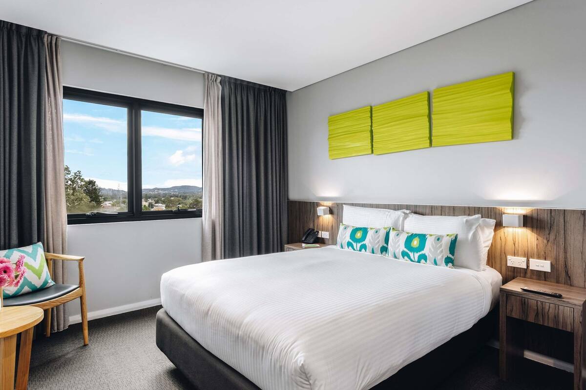 Habitación de hotel con una cama grande, un escritorio con silla, una ventana con cortinas y un aplique de pared.